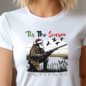 May include: White t-shirt with a green, red, and white graphic of Santa Claus hunting ducks. The text 'Tis The Season' is above the graphic.