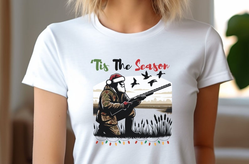 May include: White t-shirt with a green, red, and white graphic of Santa Claus hunting ducks. The text 'Tis The Season' is above the graphic.