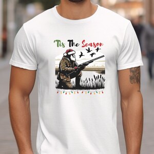 May include: White t-shirt with a Christmas-themed design featuring a Santa Claus character holding a shotgun and aiming at flying ducks. The text "'Tis The Season" is above the image.