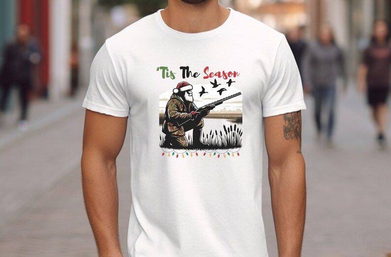 May include: White t-shirt with a Christmas-themed design featuring a Santa Claus character holding a shotgun and aiming at flying ducks. The text "'Tis The Season" is above the image.