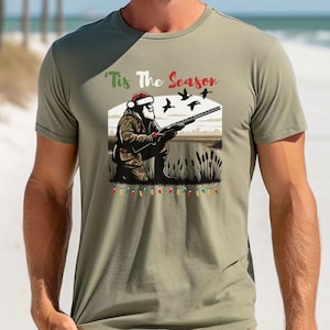 May include: A green t-shirt with a Christmas-themed design featuring a cartoon Santa Claus holding a shotgun and ducks flying overhead. The text "Tis The Season" is printed on the shirt.