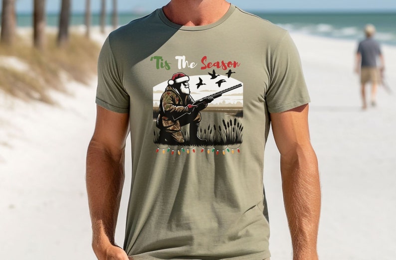 May include: A green t-shirt with a Christmas-themed design featuring a cartoon Santa Claus holding a shotgun and ducks flying overhead. The text "Tis The Season" is printed on the shirt.