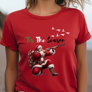 May include: Red t-shirt featuring a graphic of Santa Claus aiming a shotgun, with the text "'Tis The Season" above. Santa is wearing a red suit and carrying a sack. White birds fly across the top.