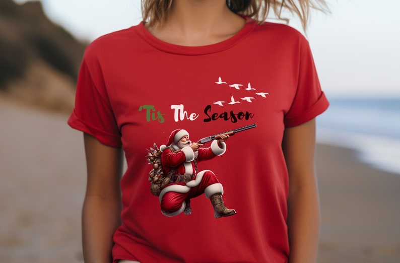 May include: Red t-shirt featuring a graphic of Santa Claus aiming a shotgun, with the text "'Tis The Season" above. Santa is wearing a red suit and carrying a sack. White birds fly across the top.