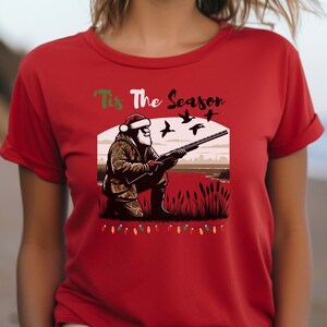May include: Red t-shirt with a Christmas-themed design featuring Santa Claus hunting ducks. The text "Tis The Season" is printed above the image.