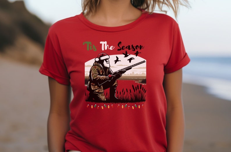 May include: Red t-shirt with a Christmas-themed design featuring Santa Claus hunting ducks. The text "Tis The Season" is printed above the image.