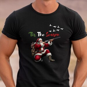 May include: Black t-shirt featuring Santa Claus aiming a shotgun, with the text "'Tis The Season" above. Santa is dressed in his traditional red suit and boots, with a sack of gifts. White birds fly in the background.