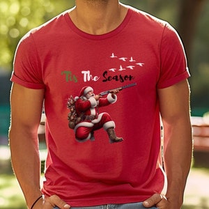 May include: Red t-shirt featuring Santa Claus aiming a shotgun at flying birds. The phrase "Tis The Season" is printed above the Santa illustration. A festive and humorous design for the holiday season.