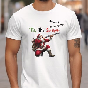 May include: White t-shirt featuring a graphic of Santa Claus aiming a shotgun at flying birds. Santa is dressed in his traditional red suit and hat. The text "'Tis The Season" is above the image.