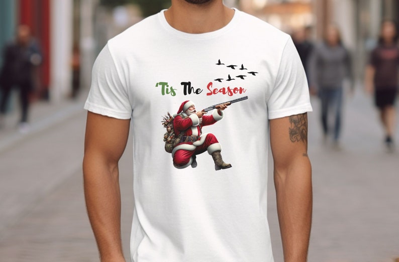 May include: White t-shirt featuring a graphic of Santa Claus aiming a shotgun at flying birds. Santa is dressed in his traditional red suit and hat. The text "'Tis The Season" is above the image.