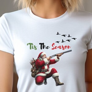May include: White t-shirt featuring a graphic of Santa Claus aiming a shotgun, with the text "'Tis The Season" above. A flock of black birds flies in the background. Santa is wearing a red suit and hat.