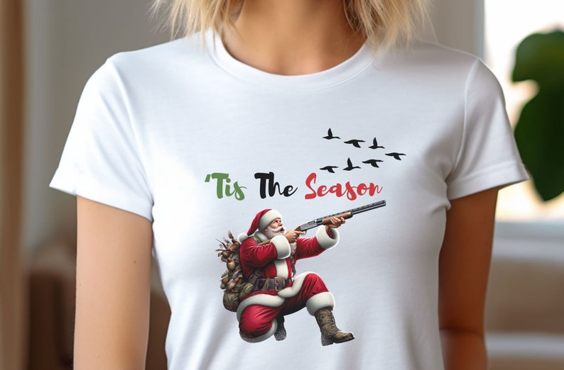 May include: White t-shirt featuring a graphic of Santa Claus aiming a shotgun, with the text "'Tis The Season" above. A flock of black birds flies in the background. Santa is wearing a red suit and hat.