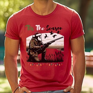 May include: Red t-shirt with a Christmas-themed design featuring Santa Claus hunting ducks. The text on the shirt reads "Tis The Season".