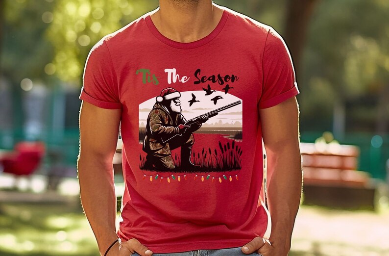 May include: Red t-shirt with a Christmas-themed design featuring Santa Claus hunting ducks. The text on the shirt reads "Tis The Season".