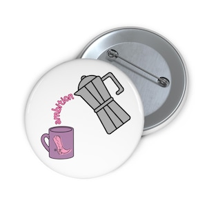Puede incluir: Una chapa redonda con fondo blanco que muestra una cafetera italiana gris vertiendo café en una taza morada clara. La taza tiene un gráfico de una bota vaquera rosa. La palabra ambition está escrita en cursiva rosa entre la cafetera y la taza.