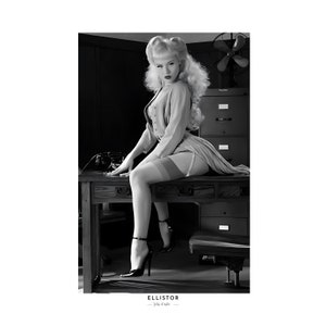 Può includere: Una fotografia in bianco e nero di una donna seduta su una scrivania in un ambiente d'ufficio vintage. Indossa una blusa bianca, una gonna e delle calze. La scrivania è in legno e ha un cassetto sul lato. C'è un ventilatore sullo sfondo.