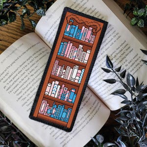 Może przedstawiać: Zakładka do książki w kształcie drewnianej biblioteczki w kolorze brązowym z wzorem haftu krzyżykowego. Biblioteczka jest wypełniona kolorowymi książkami. Zakładka jest świetnym prezentem dla miłośników książek.