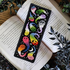 Peut inclure: Marque-page noir avec un motif pixélisé coloré de fantômes souriants et d'autres créatures fantaisistes. Le marque-page est posé sur un livre ouvert. Un accessoire unique pour les amateurs de livres.