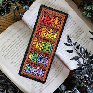 May include: A colorful bookmark with a bookshelf design featuring a variety of books with different colored spines. The bookmark is made of felt and has a brown wooden frame.