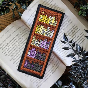 May include: A brown wooden bookshelf bookmark with a cross-stitch design. The bookshelf is filled with colourful books. The bookmark is perfect for book lovers.