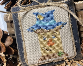 Puede incluir: Un bordado de punto de cruz hecho a mano de un espantapájaros con un sombrero azul y una cara marrón. El bordado está enmarcado en un marco de madera negro con un borde de arpillera.