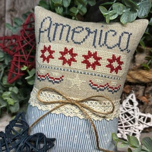 May include: A decorative pillow with the word "America" stitched in blue thread, along with red starbursts and a red, white, and blue border. The pillow has a beige top, a blue and white striped bottom, and is tied with twine.