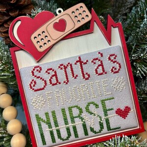 May include: A Christmas ornament with a red frame and a cross-stitched design. The design says "Santa's Favorite Nurse" in red and green. A red heart and bandage are at the top. The text "Original Design by Crafty Bluebonnet Designs" is at the top.