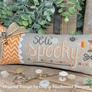 May include: A decorative pillow with the words "Sew Spooky" embroidered in orange and black. The pillow features a chevron pattern, a bow, and sewing-themed elements. A pair of scissors and buttons are in the foreground.