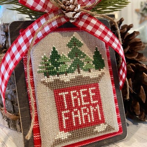 May include: A wooden sign with a red and white checkered ribbon and a pine sprig. The sign has a cross-stitched design of three green trees and the words "Tree Farm" in red.