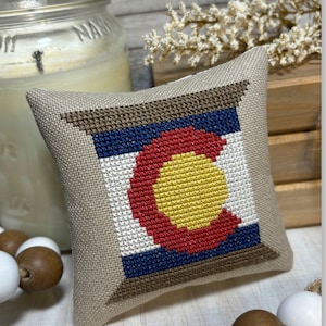 May include: A small, beige pillow with a cross-stitch design of the Colorado state flag. The flag features a large, red 'C' with a yellow circle inside, surrounded by blue and white stripes. The pillow is sitting on a wooden surface with white and brown beads.