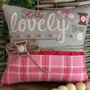 May include: A cushion embroidered with the words "Sew Lovely" in pink. It has a grey top section with cross-stitch, a red polka dot band, and a pink plaid bottom. A small thread spool is attached with twine, and the design is sewing themed.