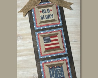 May include: A wooden wall hanging with three cross-stitched panels featuring patriotic designs. The top panel reads "OLD GLORY," the middle panel depicts the American flag, and the bottom panel displays "1776." A burlap bow and star embellish the top.