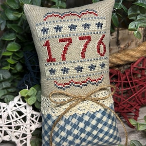 May include: A patriotic decorative pillow with the year "1776" stitched in red thread. The top portion is beige with red and blue accents, and the bottom features a blue and white checkered pattern. A lace trim and twine bow add detail.