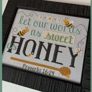 May include: Cross-stitch embroidery framed in black wood with a white background. The embroidery features a bee, flowers, and the text "Let our words be as sweet as honey" with the scripture reference "Proverbs 16:24".