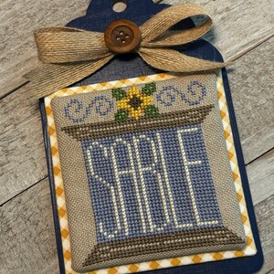 May include: A blue and brown cross-stitch design with the word "SALE" in white lettering. The design is framed with a yellow and white checkered border and a sunflower is at the top. The design is mounted on a blue wooden tag with a brown button and a burlap bow.