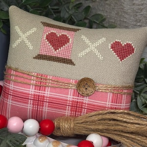 May include: A beige pillow with a red heart and the letters "XO" embroidered on it. The pillow has a pink and white plaid fabric on the bottom and a brown button. The pillow is sitting on a white surface with a string of pink, white, and red beads.