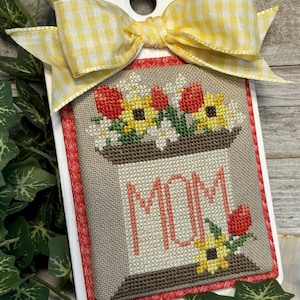 May include: A handmade cross-stitch piece with a floral design and the word "MOM". The piece is framed in a white wooden plaque with a yellow and white gingham bow at the top. The design includes red tulips and yellow sunflowers.