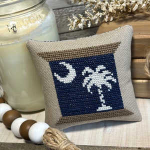 May include: A small, beige pillow with a brown frame and a blue background. The pillow features a white embroidered palm tree and crescent moon, representing the state of South Carolina.