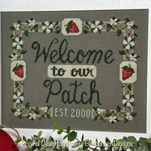 Puede incluir: Obra de arte en punto de cruz enmarcada con las palabras "Welcome to our Patch" y "EST. 2000". El diseño presenta fresas y detalles florales en un marco blanco. El fondo es gris apagado.