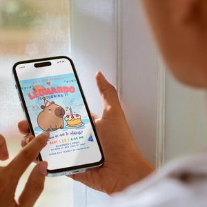 May include: A smartphone displaying a birthday invitation for Leonardo's 1st birthday party. The invitation features a cartoon illustration of a capybara wearing a party hat and holding a slice of cake. The text on the invitation reads "It's a Capy Birthday! Leonardo is turning 1! Join us as we celebrate! Sept 1, 2:00-4:00 PM Alexander's House 945 Main St, New York, NY Please RSVP to [email protected] by [date]"