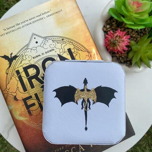 May include: A light blue square case with a black and gold glitter dragon design. The case is on top of a book with the title "Iron Flame" by Rebecca Yarros. A small succulent plant is in the background.