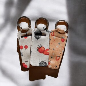 May include: Three keyrings with different fabric patterns. The keyrings are white with black and red accents, white with black and red accents, and beige with red accents. The keyrings have a silver ring.