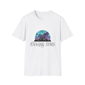 Op de afbeelding: Wit T-shirt met een hemels ontwerp. De afbeelding toont een draaiende melkwegscène in turquoise en paars boven een zwarte bergketen. De woorden "Chasing Stars" staan eronder in een sierlijk lettertype.
