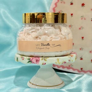 May include: A jar of warm vanilla sugar whipped soap with a gold lid, displayed on a small, decorative, floral-patterned cake stand. The label on the jar reads "Warm Vanilla Sugar Whipped Soap."