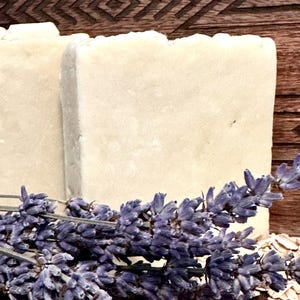 May include: Two rectangular bars of light-colored soap are displayed with sprigs of purple lavender and scattered oats. The soap has a rough, textured appearance. The background is a dark wooden surface with a carved geometric design.