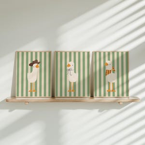 May include: Three framed prints with green and white striped backgrounds and illustrations of geese. The geese are wearing different accessories, including a cowboy hat, a bow, and a scarf.