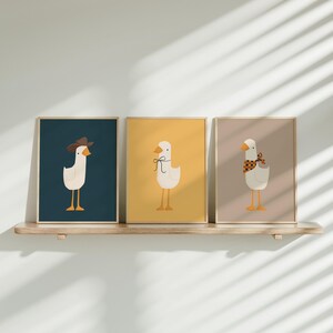 May include: Three framed illustrations of geese with different accessories. The first goose is wearing a cowboy hat, the second goose is wearing a bow tie, and the third goose is wearing a bandana. The frames are all different colors: blue, yellow, and beige.