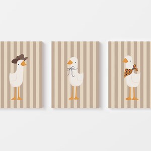 May include: Three printable wall art prints featuring a white goose with different accessories. The first goose is wearing a brown cowboy hat, the second goose is wearing a black and orange bow, and the third goose is wearing a red and black checkered scarf. The background of each print is a light brown with vertical stripes.