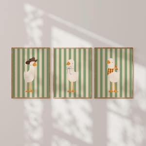 May include: Three framed prints with a green and white striped background. Each print features a white goose with different accessories. The first goose is wearing a brown cowboy hat, the second goose is wearing a brown bow tie, and the third goose is wearing a plaid scarf.