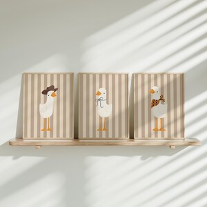 May include: Three framed prints with a striped background and illustrations of geese. The geese are wearing different accessories, including a cowboy hat, a bow, and a scarf.